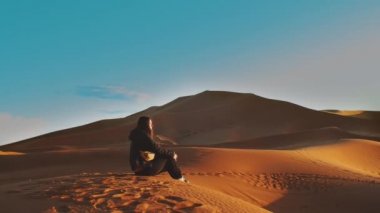 Sahra çölünde gün batımında kumullarda yalnız başına oturan uzun saçlı bir kadın. Turist, sonsuz çölde kumlu bir tepenin üzerinde tek başına oturuyor. Güzel manzaralar, Fas, tam kapasite.