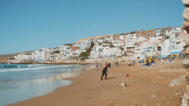 Muitas pessoas relaxando na praia em pequena aldeia, fundo do oceano. Meninos brincando com bola rosa, gaivotas voam sobre, Marrocos, hd completo — Vídeo de Stock