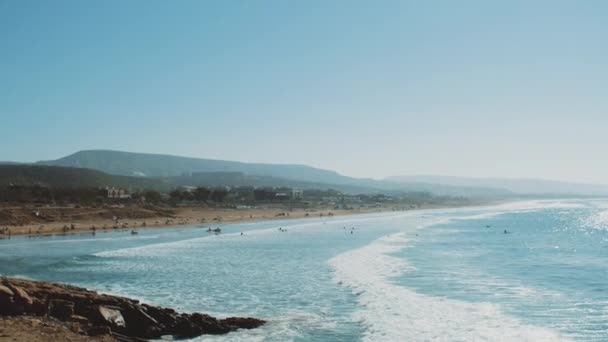 Surfing i Atlanten, Marocko, många människor på stranden, stora vågor och klar himmel och berg vid horisonten, Taghazout byn, full hd — Stockvideo