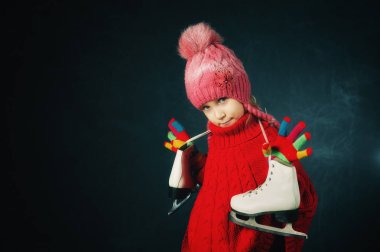 Stüdyo bir kırmızı kazak ve örme şapka koyu arka plan üzerinde küçük bir kız portresi. Kız holding paten