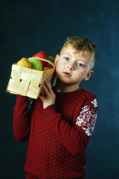 Boy holding apples on dark background . The child is dressed in a warm sweater, holding a fruit basket