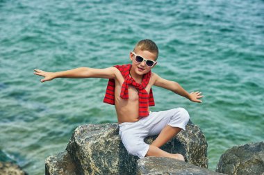 Deniz kıyısında moda yakışıklı çocuk . Dinlenme ve seyahat . Çocuk hafif pantolon bir taş üzerinde oturur .