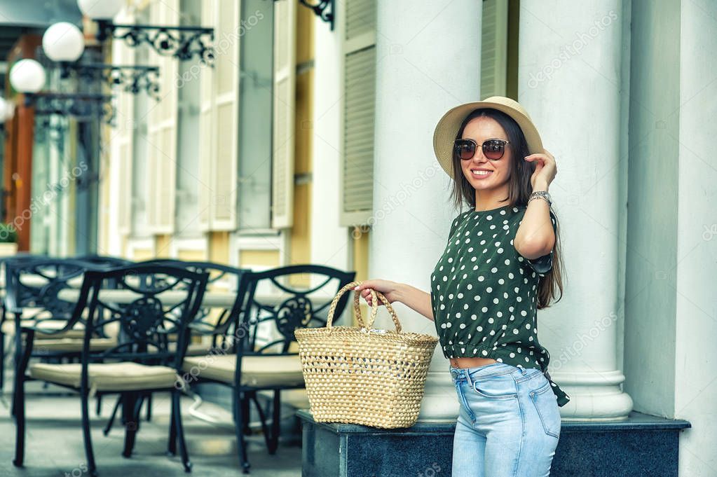 Young woman in casual clothes in summer wide-brimmed hat and with vintage braided bag on the city street .