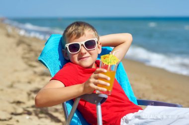 Çocuk deniz kıyısında dinleniyor. Bir bardak meyve suyu tutan bir plaj sandalyesinde oturan çocuk. Çocuk eğlencesi kavramı