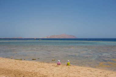 İki sevimli küçük bebek deniz suyu yakınındaki kum plajındaki otur. Oğlan ve kız şapka ve t-shirt giyiyor. Yatay renkli fotoğraf.