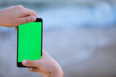 İki küçük elleri boş boş yeşil ekran monitör ile modern siyah smartphone tutan genç çocuğun görünümünü closeup bulanık mavi deniz su arka plan üzerinde izole. Yatay fotoğraf ile boşaltmak.