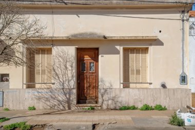 Ahşap panjurları, merkezi kapısı ve güneş ışığıyla aydınlanmış gölgesi olan alçak, yaşlı bir banliyö evi..