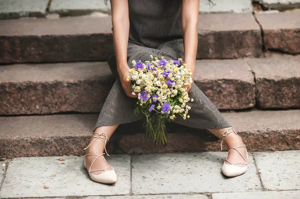 a girl in a light dress sits on the steps and holds a bouquet of wild flowers in her hands