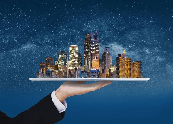 Smart city, smart building, real estate business and investment. Businessman hand holding digital tablet with modern building on blue background and starry sky