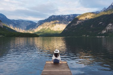 Ahşap iskelede tek başına oturan genç kadın, yaz aylarında Hallstatt, Avusturya'da göl ve dağ manzarasına bakıyor