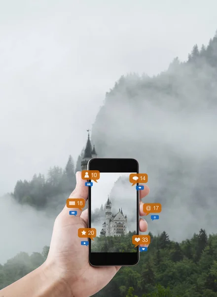 ソーシャルメディアやソーシャルネットワークの通知アイコンで ドイツのノイシュヴァンシュタイン城の写真を撮る携帯電話を使用した手 — ストック写真