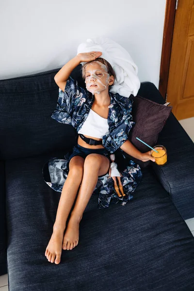 Маленькая девочка, как мама, в косметической маске на лице, сидит на диване с коктейлем в отпуске Лицензионные Стоковые Фото