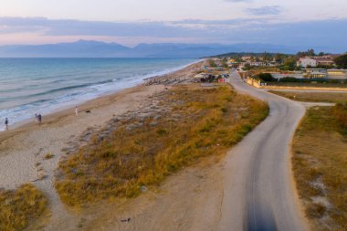 Serene Ionian Coastline at Sunset: An aerial view capturing the tranquil beauty of a Greek beach with gentle waves, distant mountains, and a peaceful road as dusk settles.