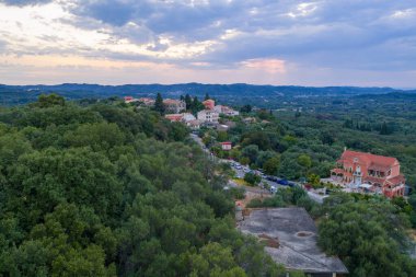 Aerial drone view of a traditional Mediterranean village in Corfu, Greece, with colorful houses, stone rooftops, winding roads, lush green hills and surrounding countryside landscape under summer sky.