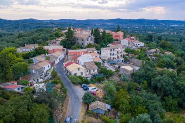 Aerial drone view of a traditional Mediterranean village in Corfu, Greece, with colorful houses, stone rooftops, winding roads, lush green hills and surrounding countryside landscape under summer sky.