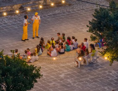 Corfu, Greece - August 17, 2025: Community Gathering in a Greek Village Square: An aerial view of a lively evening event in a traditional village square, with a crowd of children sitting on the ground watching a performance by two entertainers under 
