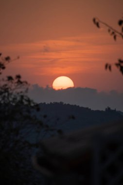 Yunanistan 'daki yuvarlanan tepeler ve dağların üzerinde büyüleyici bir günbatımı. Güneş kısmen bulutlarla kararmış, sıcak turuncu bir parıltı yayıyor..