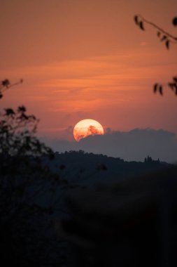 Yunanistan 'daki yuvarlanan tepeler ve dağların üzerinde büyüleyici bir günbatımı. Güneş kısmen bulutlarla kararmış, sıcak turuncu bir parıltı yayıyor..