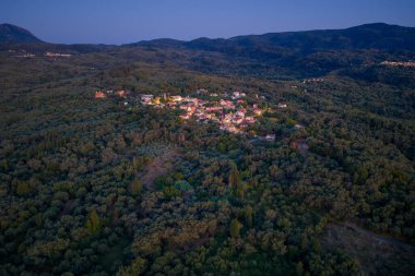 Alacakaranlıktaki Bereketli Ormandaki Dağ Köyü: Küçük, geleneksel bir köyün havadan görünüşü akşam karanlığının ilk ışıklarıyla yeşil bir ormana yerleşmiş.