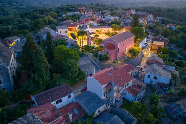 Alacakaranlıktaki Büyüleyici Yunan Köyü: Alacakaranlıkta sokak lambaları parıldamaya başladığında yemyeşil bir ormanda yuva yapmış küçük, geleneksel bir köyün havadan görünüşü sakin ve huzurlu atmosferi vurguluyor.