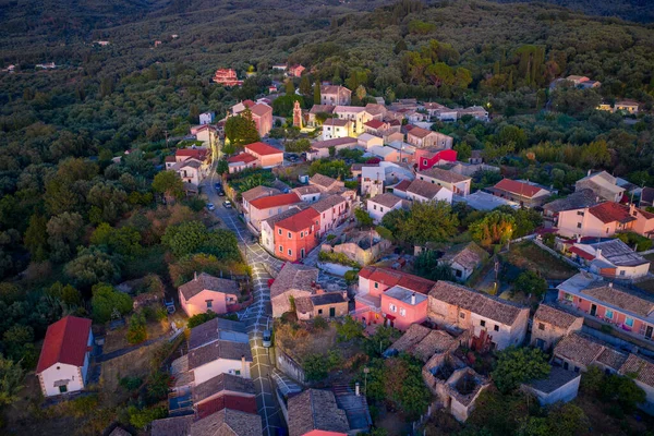 Alacakaranlıktaki Büyüleyici Yunan Köyü: Alacakaranlıkta sokak lambaları parıldamaya başladığında yemyeşil bir ormanda yuva yapmış küçük, geleneksel bir köyün havadan görünüşü sakin ve huzurlu atmosferi vurguluyor.