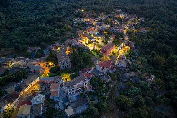 Alacakaranlıktaki Büyüleyici Yunan Köyü: Alacakaranlıkta sokak lambaları parıldamaya başladığında yemyeşil bir ormanda yuva yapmış küçük, geleneksel bir köyün havadan görünüşü sakin ve huzurlu atmosferi vurguluyor.