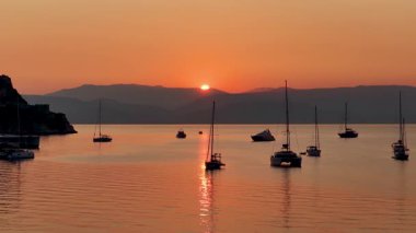 Sunrise over Corfu harbor in Greece with yachts and sailboats anchored on calm sea waters, glowing orange horizon, Old Fortress silhouette and tranquil Mediterranean seascape, idyllic summer travel des