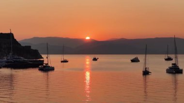 Sunrise over Corfu harbor in Greece with yachts and sailboats anchored on calm sea waters, glowing orange horizon, Old Fortress silhouette and tranquil Mediterranean seascape, idyllic summer travel des