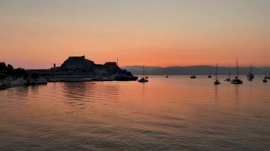Aerial drone view of the Old Fortress in Corfu Town, Greece, at sunrise. Sailboats and yachts are anchored in the calm Ionian sea against a beautiful orange and pink sky during the golden hour.