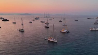 Stunning aerial drone panorama of luxury yachts, catamarans, and sailboats anchored in a calm Mediterranean bay in Greece. A beautiful, peaceful seascape at sunset with a pink and orange sky.