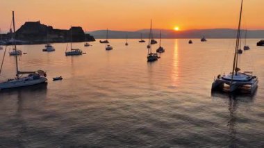 Sunrise over Corfu harbor in Greece with yachts and sailboats anchored on calm sea waters, glowing orange horizon, Old Fortress silhouette and tranquil Mediterranean seascape, idyllic summer travel des