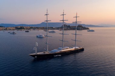 Majestic three-mast luxury superyacht anchored in a tranquil sea at sunrise. Aerial drone view of the modern vessel against a beautiful orange sky and a hazy, mountainous coastline.
