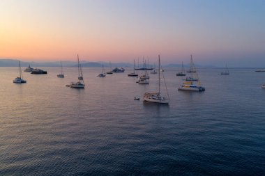 Captivating aerial view of numerous sailboats and yachts anchored in the tranquil sea during a serene sunset, with distant mountains silhouetted under a pastel sky.