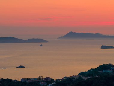 Yunanistan 'ın Korfu kıyısında, alacakaranlıkta sakin bir deniz manzarası. Sakin İyon Denizi siluetli adaların arkasındaki canlı turuncu ve pembe gün batımını yansıtır..