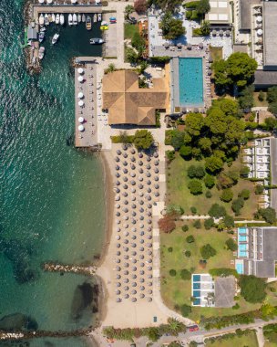 Yunanistan 'ın Korfu kentinde lüks bir sahil otelinin insansız hava aracı perspektifi. Resimde tatil köyünün havuzu, özel yat limanı ve sayısız güneş şemsiyesi bulunan kumlu sahil görülüyor.