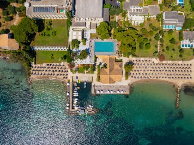 Yunanistan 'daki lüks bir otel otelinin tepeden tırnağa insansız hava aracı görüntüsü. Yüzme havuzunda dinlenen insanlar, özel bir tekne iskelesi ve Korfu sahilinde şemsiyeli kumlu bir sahil.