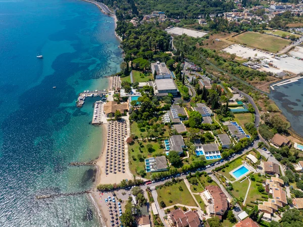 Korfu 'daki bir Yunan adası tatil beldesinin yüksek açılı insansız hava aracı görüntüsü. Sahile yakın bir otel, yüzme havuzları, küçük bir tekne iskelesi ve İyon Se 'nin geniş mavi suları yer alıyor.
