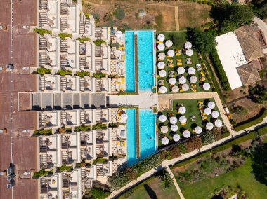 Yunanistan 'ın Corfu kentindeki lüks bir tatil beldesinin yukarıdan aşağıya insansız hava aracı görüntüsü. Otel binasının simetrik tasarımı, havuzlar ve düzgün düzenlenmiş güneş yatakları çarpıcı bir desen oluşturuyor.
