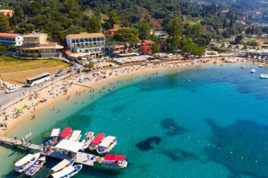 Aerial drone view of a popular tourist beach in Greece with beautiful turquoise water. People swimming and relaxing on the sand next to a pier with boats and waterfront hotels and restaurants