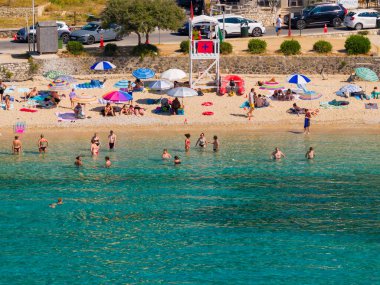 A lively public beach in Greece on a sunny day. People with colourful umbrellas relax on the sand while families play and swim in the beautiful clear turquoise water near a lifeguard tower