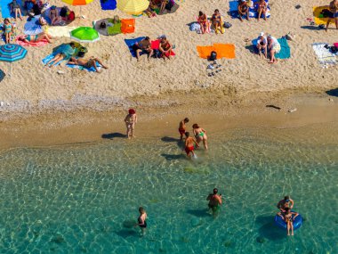 Top-down view of people sunbathing and swimming at a beach in Greece. Tourists relax on colourful towels on the sand and enjoy the crystal-clear turquoise sea