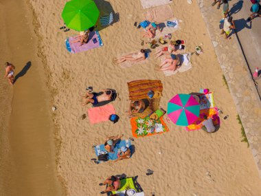 Birçok insanın yaz tatilinin keyfini çıkardığı, havluların üzerinde dinlendiği ve sıcak altın kumda renkli şemsiyeler altında geçen canlı bir sahil manzarasının insansız hava aracı görüntüsü.