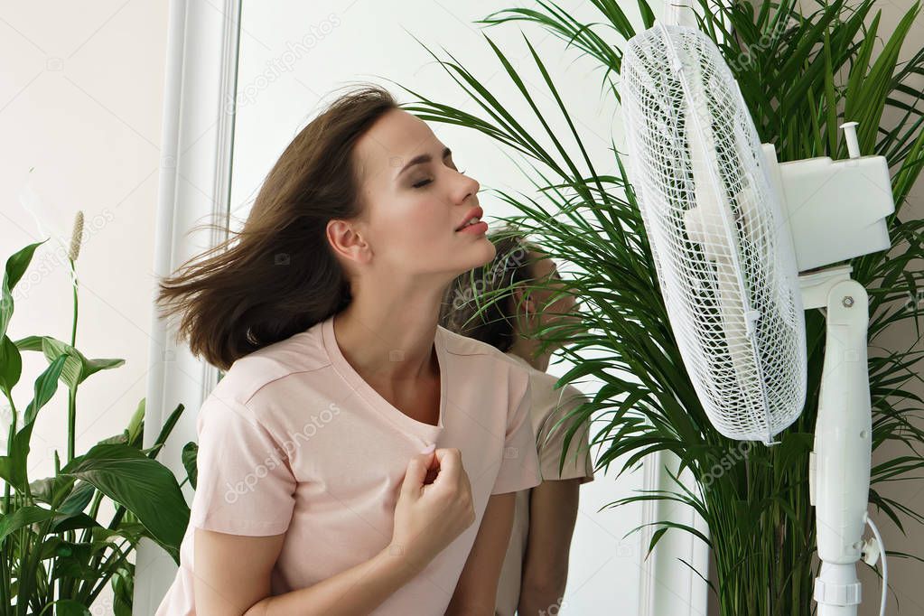 Young woman cooling herself by electric fan at home 