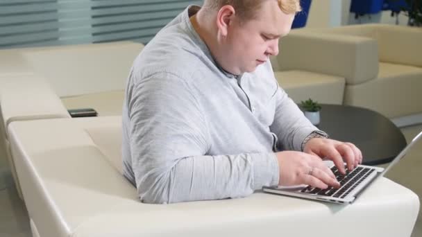 Concepto de trabajo de oficina. El hombre está navegando en su computadora portátil, sentado en la oficina en el acogedor sofá blanco — Vídeos de Stock