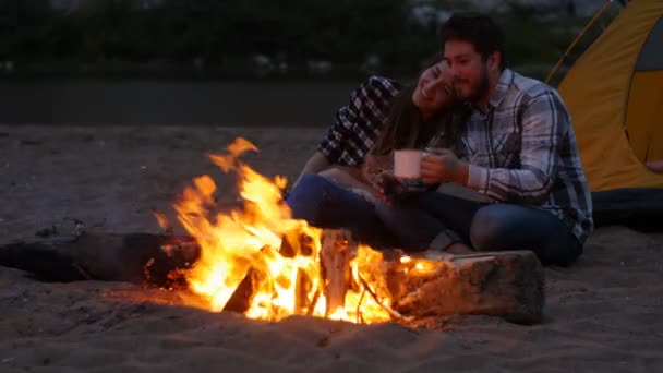 Koncepció, a barátság, a szeretet és a kapcsolatok. Fiatal boldog pár ül a strandon, este tábortűz — Stock videók
