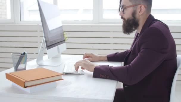 Концепция офисной работы или фриланса. Молодой бородатый стильный мужчина работает за компьютером в офисе — стоковое видео