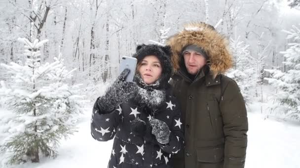 Concepto de viaje de invierno. Feliz pareja joven haciendo foto selfie en el teléfono inteligente al aire libre con bosque de nieve en el fondo — Vídeo de stock