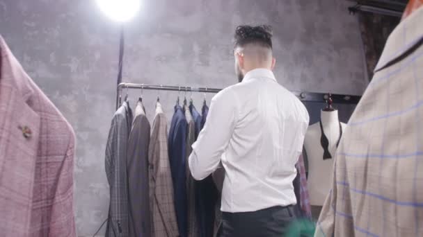 Концепция шопинга и моды - молодой бородатый мужчина выбирает и примеряет куртку в магазине одежды — стоковое видео