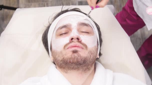Концепція спа для чоловіків. Молодий чоловік наносить косметичну маску на обличчя в салоні краси — стокове відео