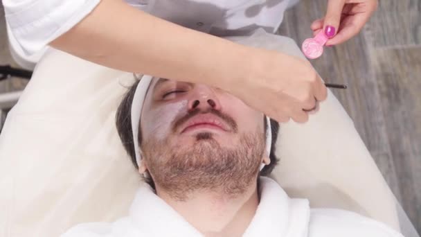 Begreppet spa för män. En ung man tillämpas en kosmetiska mask i ansiktet i en skönhetssalong — Stockvideo